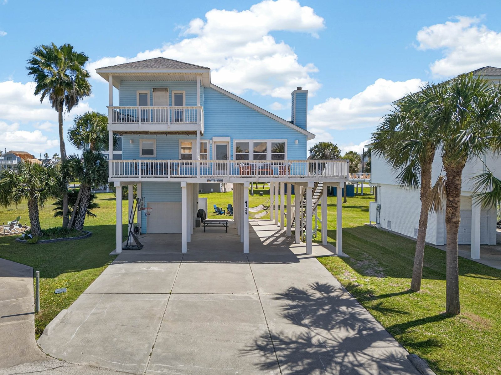 Front exterior of a two-story light-blue elevated beach house with balconies, palm trees and covered parking at Salty Sea + Sunlight.